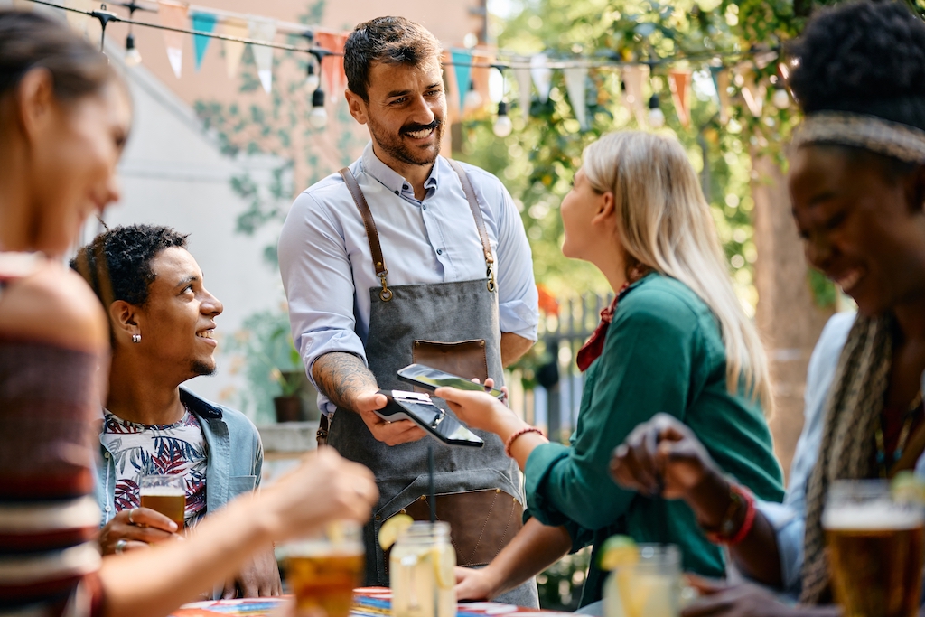 Happy waiter receiving smart phone contactless payment form a woman who is being with her friends in a cafe.